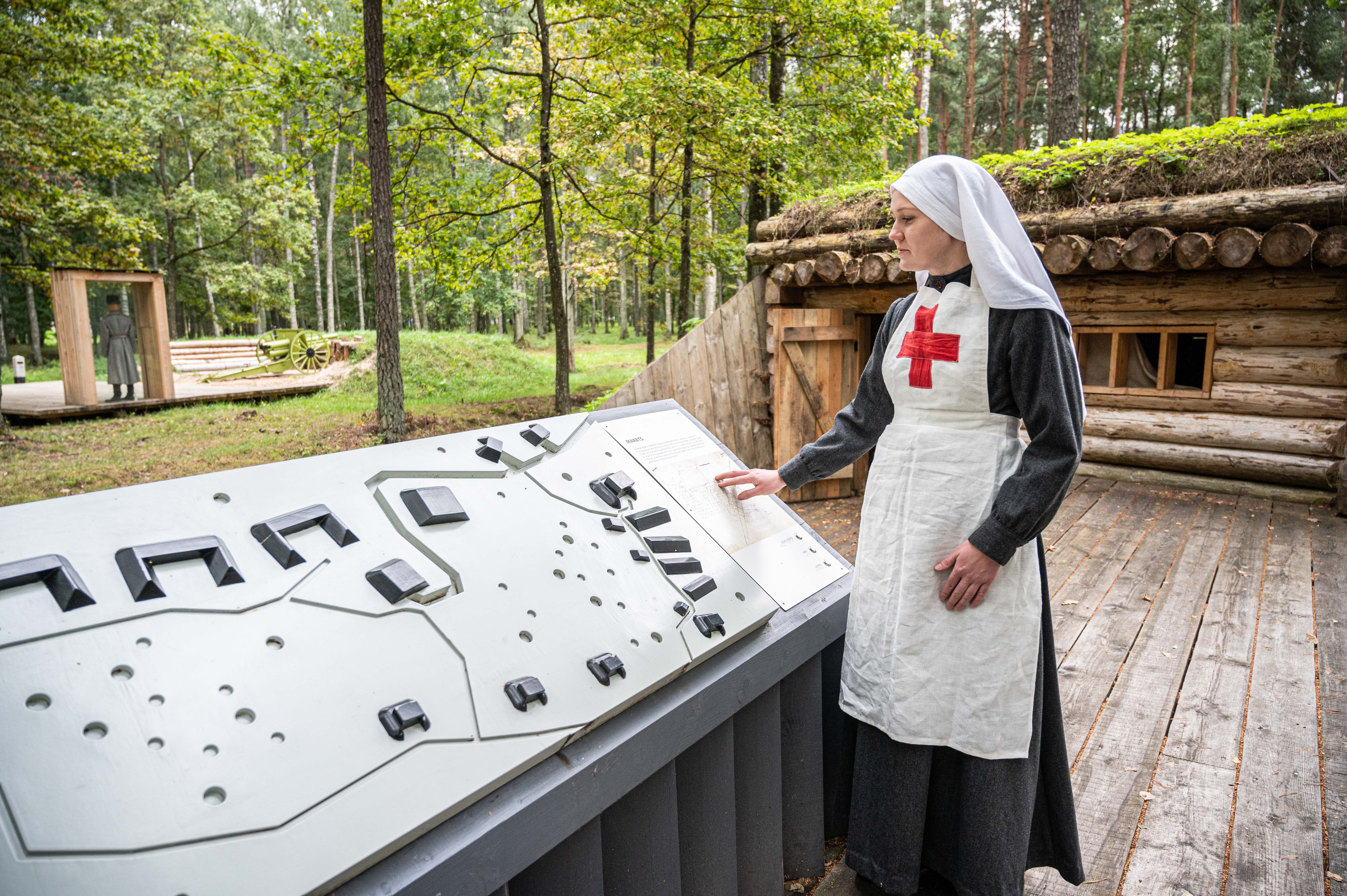 Priekšplānā redzams taktilais makets un sieviete Pirmā pasaules kara medmāsas tērpā. Aizmugurē redzama zemnīca.  